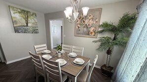 Un coin repas avec une table ronde, six chaises et un lustre. Deux tableaux ornent les murs, une porte se trouve au fond, et une plante en pot est placée à droite.
