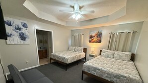 Une chambre avec deux lits, un ventilateur de plafond, une table de chevet avec une lampe et un banc gris.