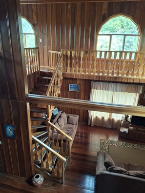 Un intérieur en bois avec une chambre à coucher située en mezzanine, un escalier et un espace de vie comprenant un canapé et un téléviseur.
