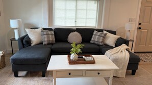 A living room with a dark-colored sectional sofa, a white coffee table, and a window with blinds. - NEW Charming 2 bdrm King and Queen (North Charleston)