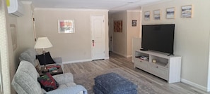 A living room with a sofa, ottoman, TV, and a door leading to another room. - 2 Bdr 2 Ba House #40 Borrego Springs CA 55+ Community golf, pool, pball, club (Borrego Springs)
