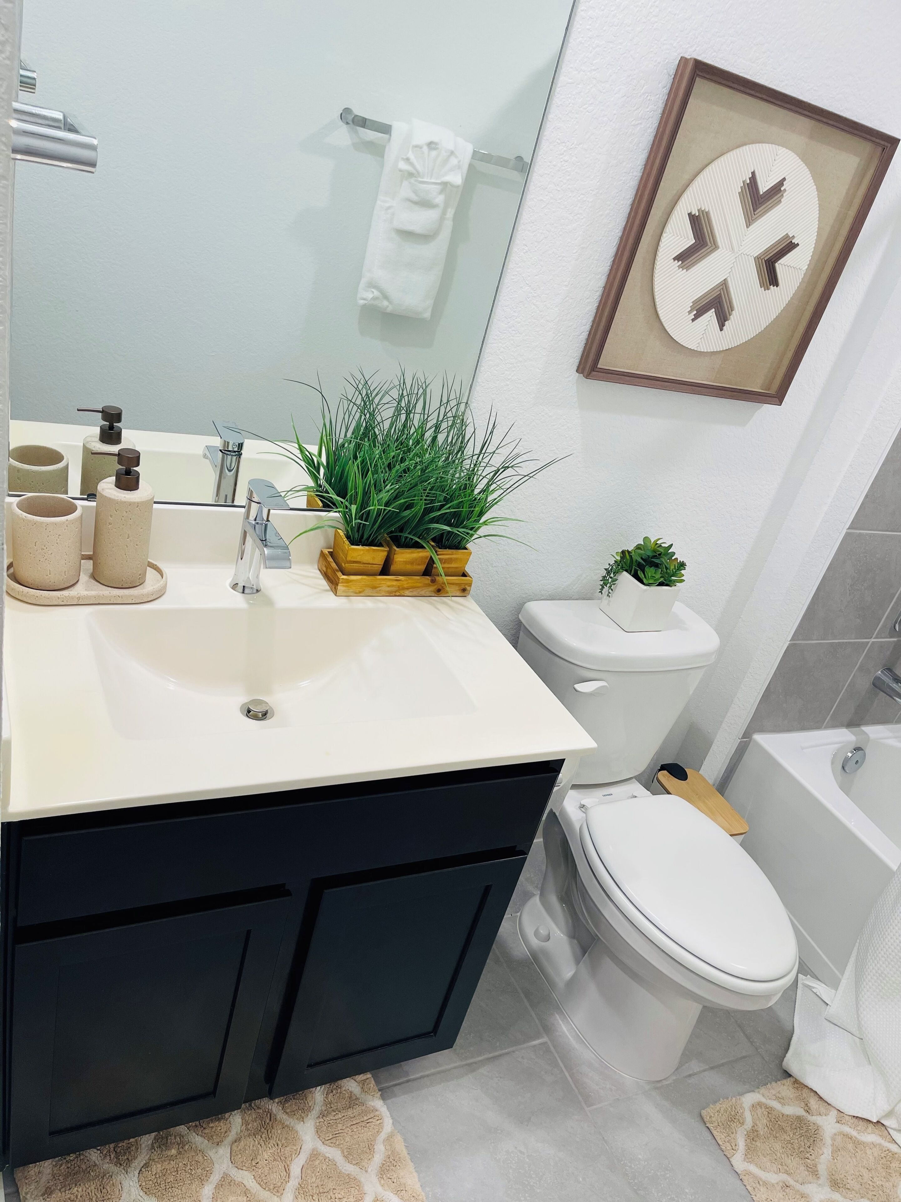 Une salle de bain avec un lavabo blanc, des meubles noirs, des toilettes et une baignoire. On peut voir une petite plante sur le lavabo et un tableau encadré au mur.