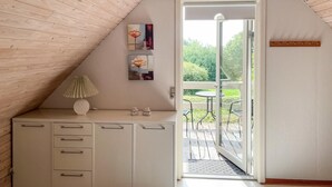 A room with a sloped ceiling, a white cabinet with drawers, a lamp, and a small table. There are two paintings on the wall, and a door leading to an outdoor patio with a table and chairs. - Lovely home in Tranekær with WiFi (Tranekær)