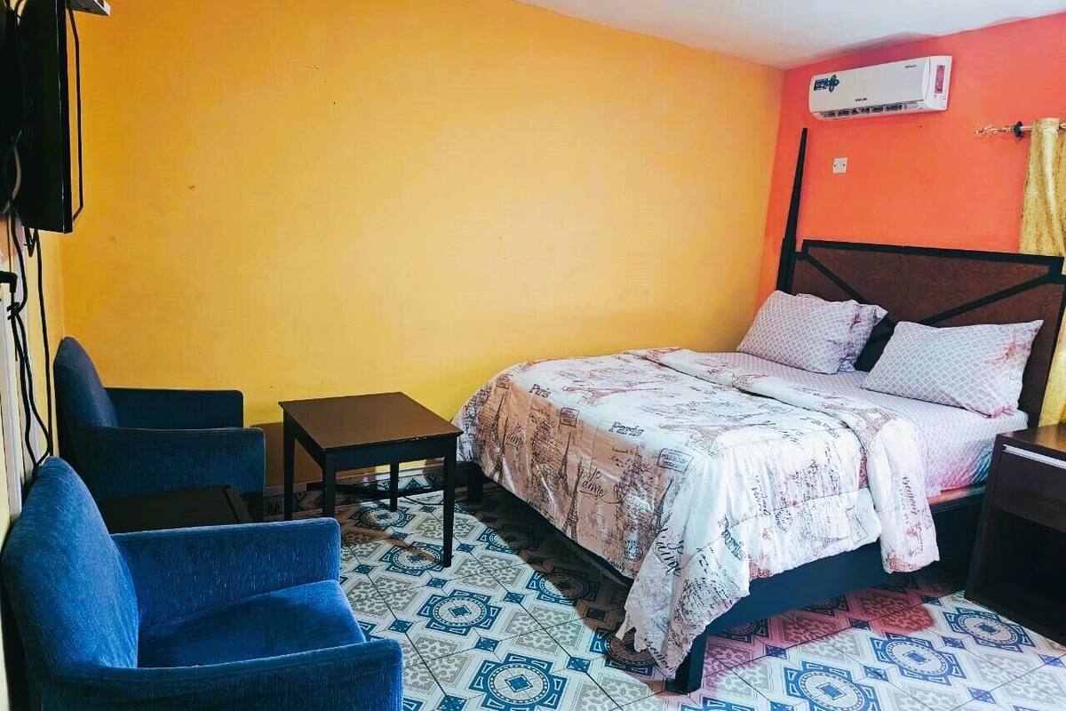 A bedroom with a bed, two blue chairs, a small table, and an air conditioning unit.