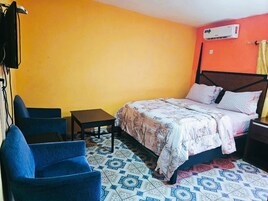 A bedroom with a bed, two blue chairs, a small table, and an air conditioning unit.