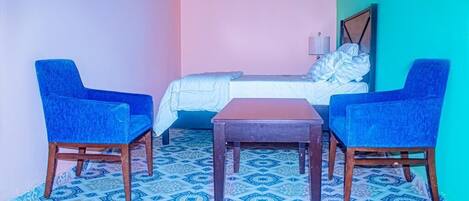 A small room with a bed, two blue chairs, a wooden table, and patterned floor tiles.