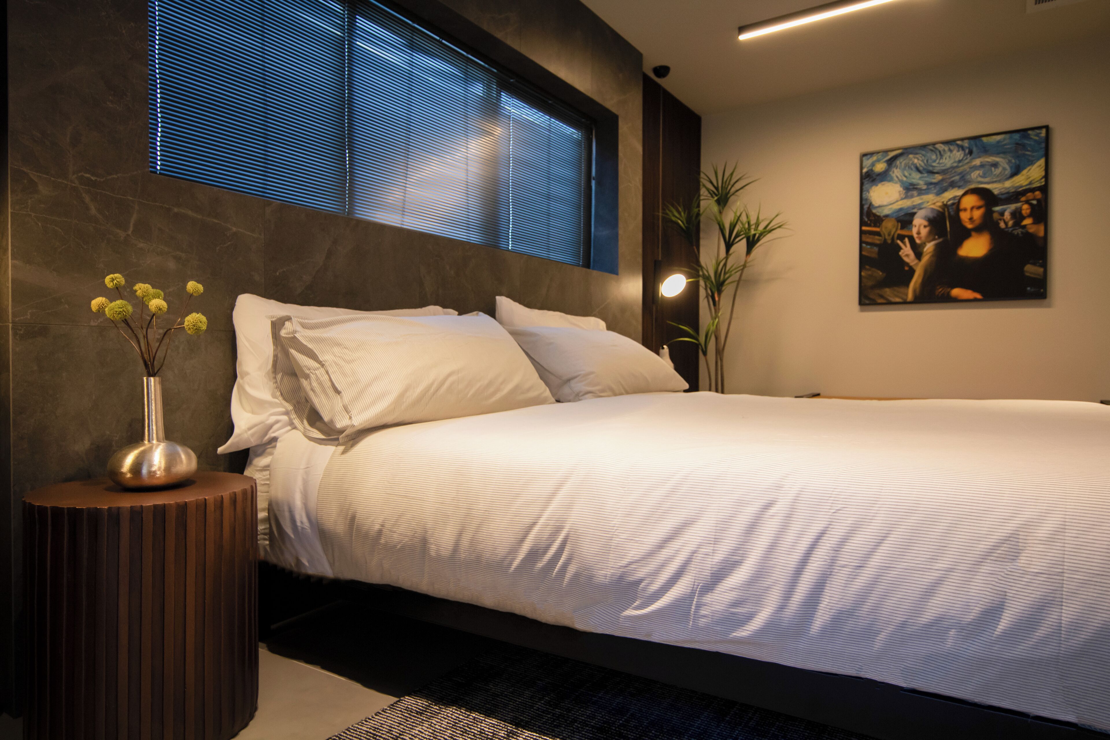 Una camera da letto moderna con un letto grande, un comodino con un vaso di fiori, un quadro appeso al muro e una finestra con persiane.
