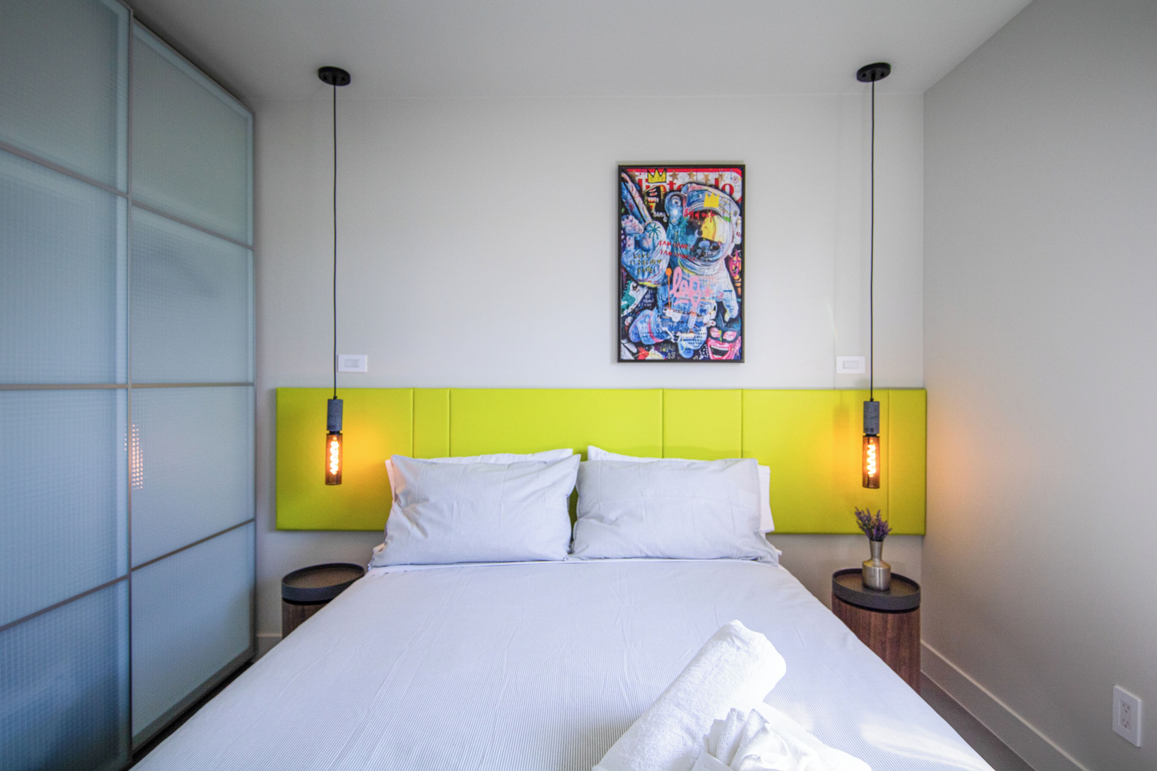 A hotel room with a bed, a green headboard, two bedside lamps, and a colorful artwork on the wall.