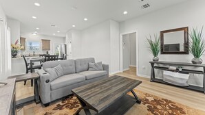 Amplia sala de estar con un sofá seccional gris, una mesa de centro de madera y un tapete estampado. Se ve un comedor con mesa y sillas al fondo.