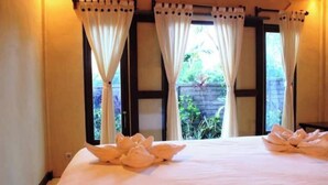 A hotel room with a large bed, white curtains, and a view of greenery outside.