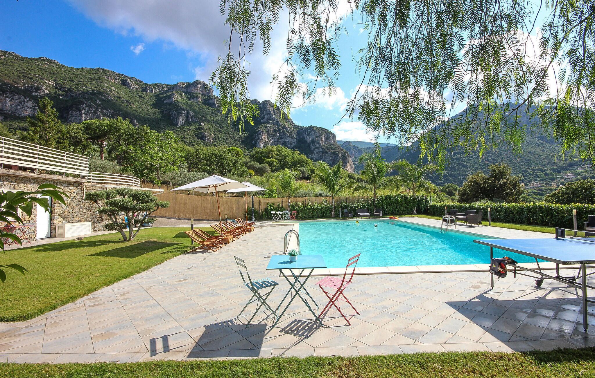 A swimming pool with lounge chairs and a table, surrounded by a stone patio and lush greenery, with mountains in the background.