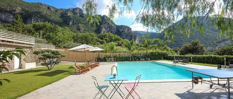 A swimming pool with lounge chairs and a table, surrounded by a stone patio and lush greenery, with mountains in the background.