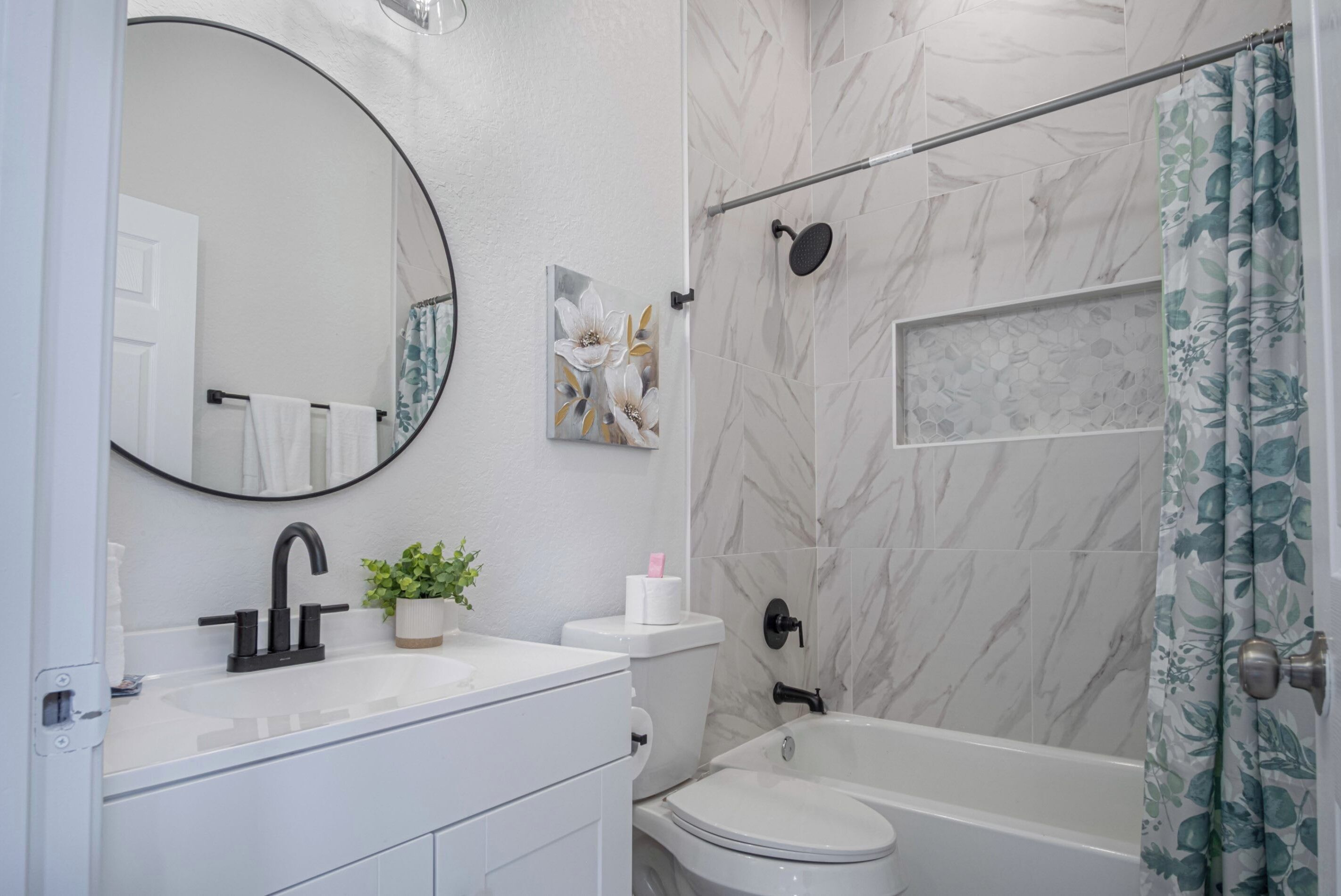 A bathroom with a white sink, black faucet, toilet, and bathtub. There is a mirror above the sink and a shower curtain with a floral pattern.