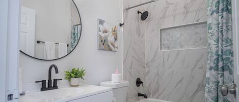 Un baño con lavamanos blanco, grifo negro, inodoro y bañera. Hay un espejo sobre el lavamanos y una cortina de ducha con motivos florales.