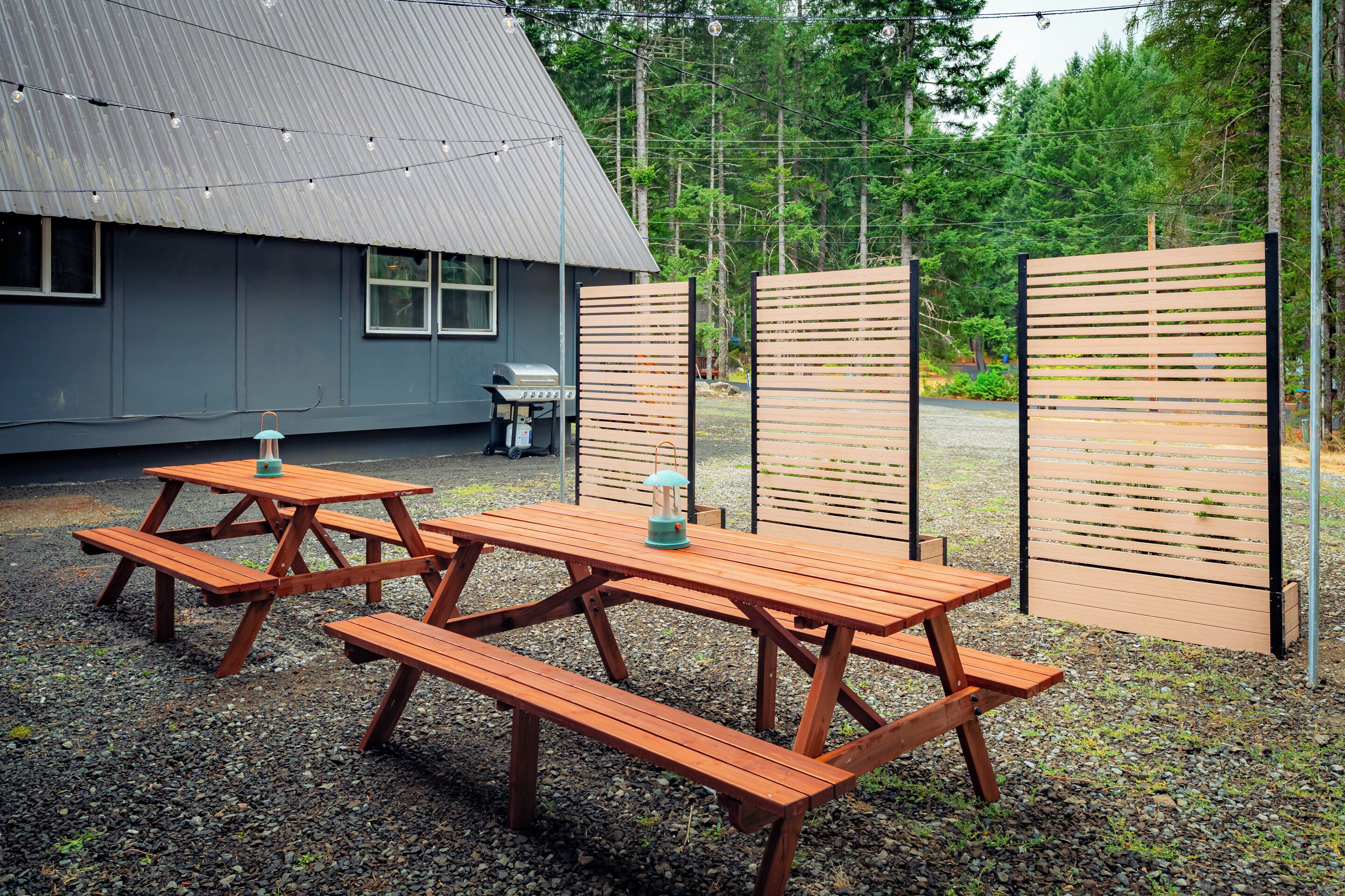 A picnic area with wooden benches and tables, a barbecue grill, and a house in the background.