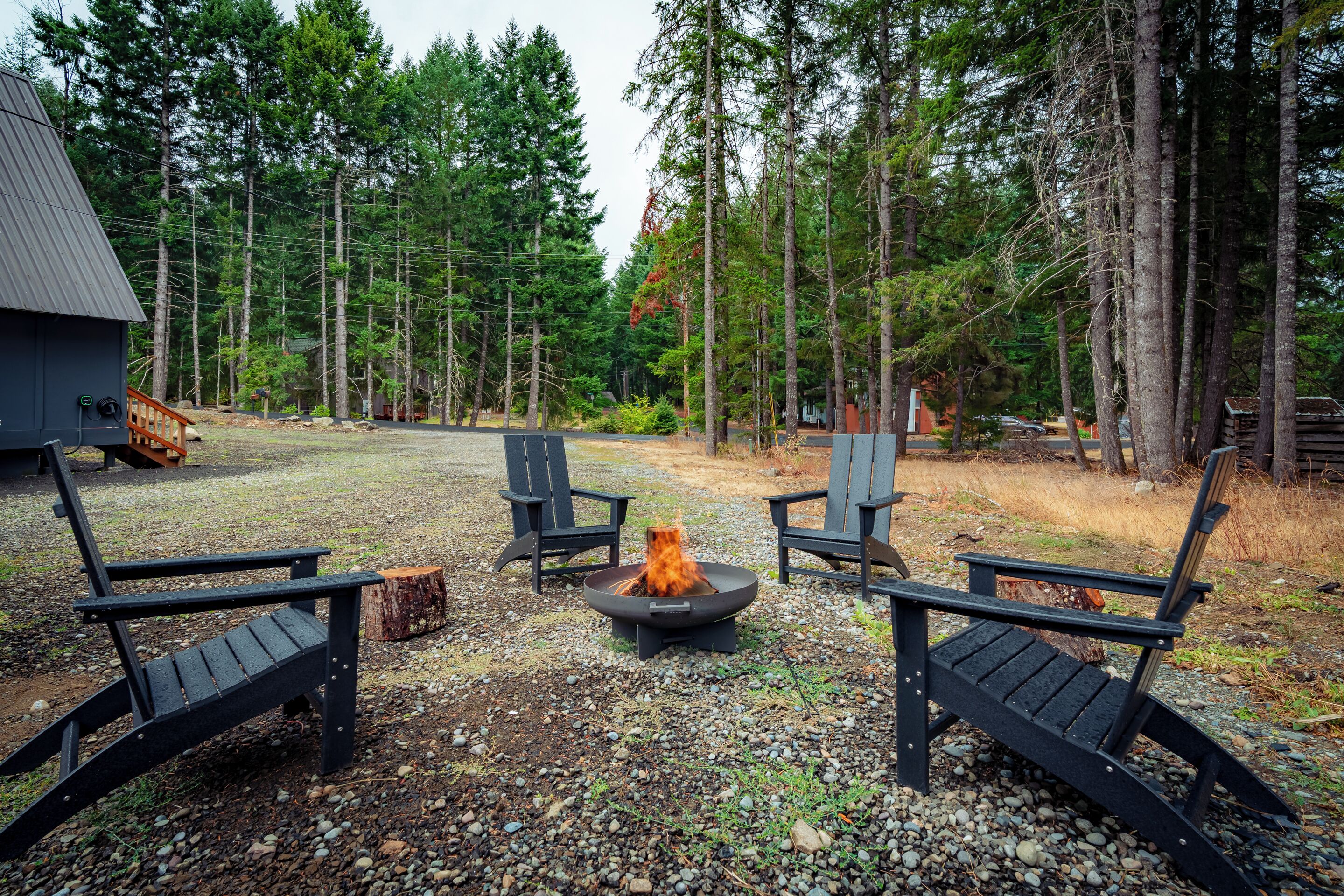 A rustic outdoor seating area with Adirondack chairs around a fire pit, surrounded by a forested landscape.