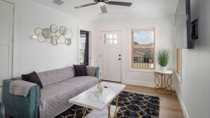 A living room with a teal sofa, a white coffee table, a ceiling fan, a door, and a window with a view of a house. - Stylish Stay With Unforgettable Downtown Views! (San Antonio)