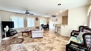 Uma sala de estar com televisão, sofá branco, mesa de jantar com cadeiras, área de cozinha com pia e banquetas, e ventilador de teto.