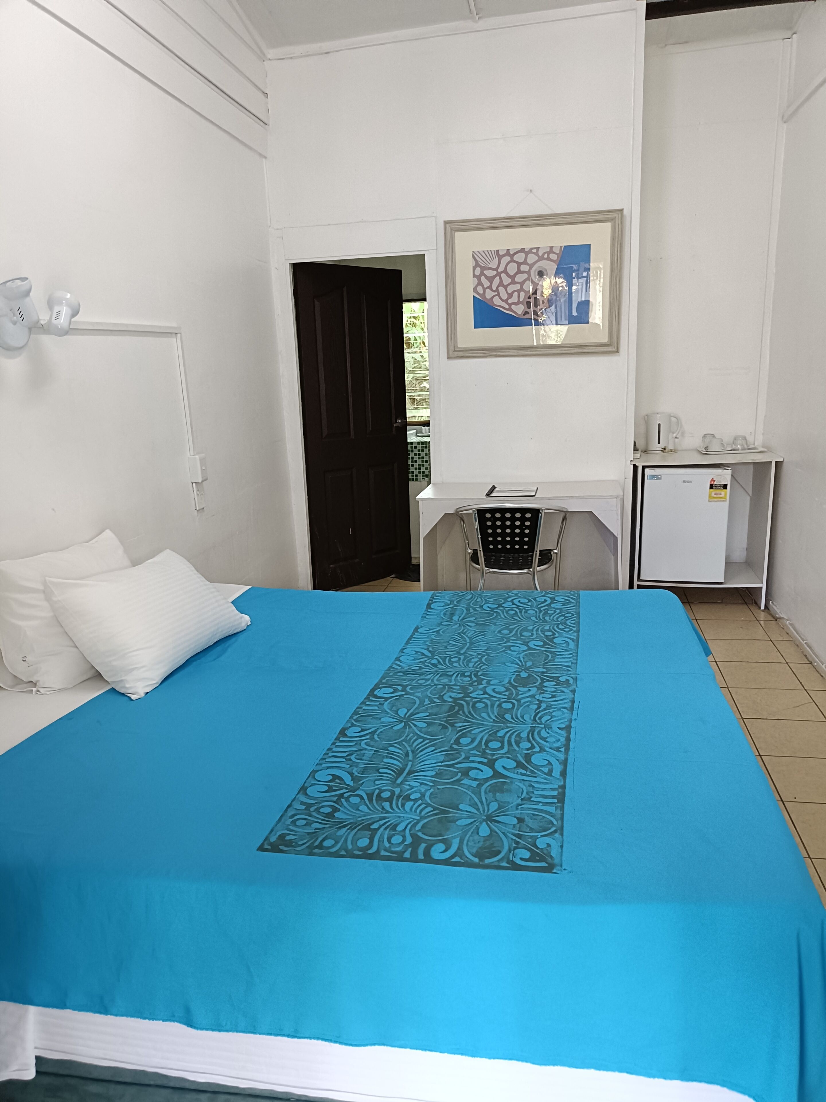 Una habitación con cama, escritorio, silla, refrigerador y puerta que da al exterior.