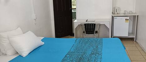Una habitación con cama, escritorio, silla, refrigerador y puerta que da al exterior.