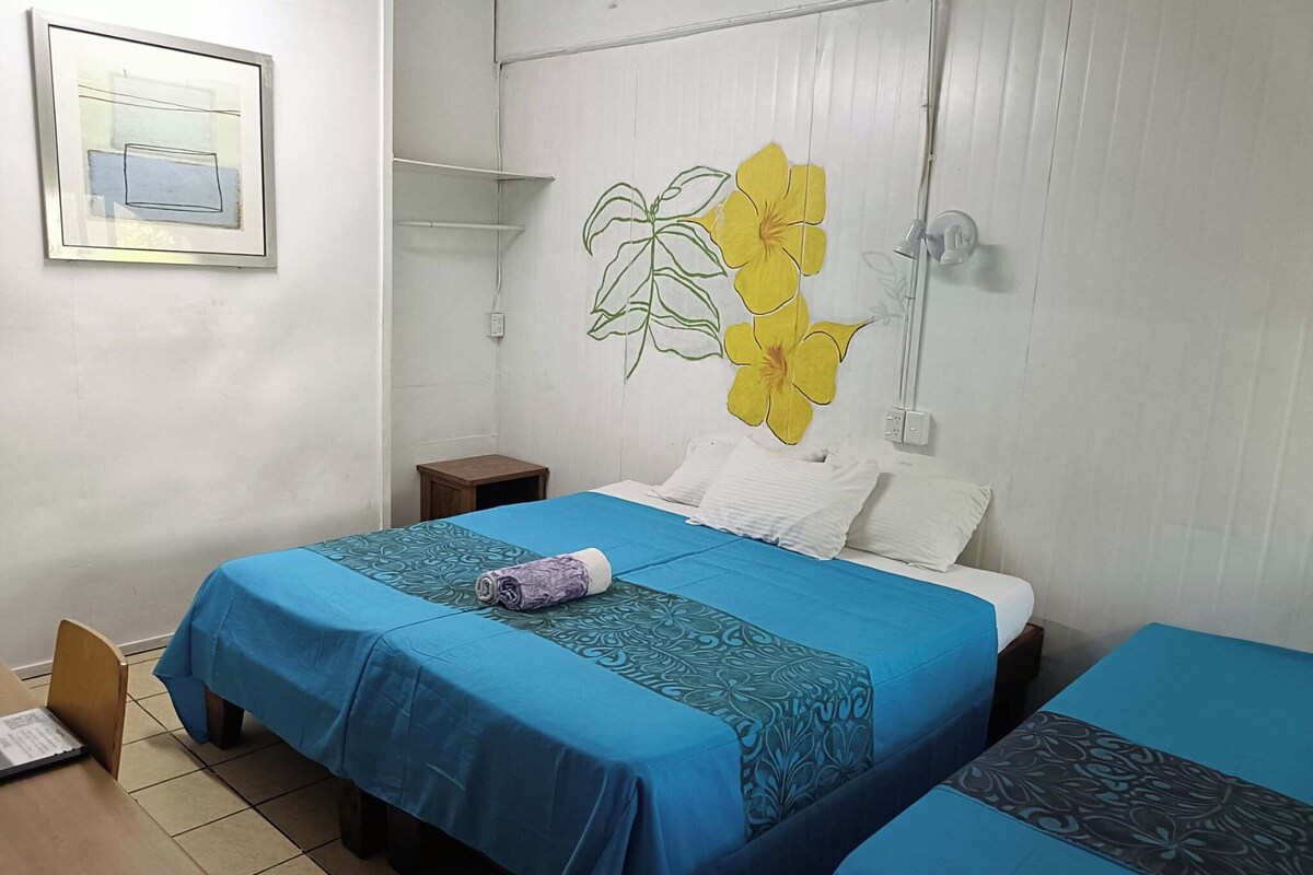 A room with two beds, a small table, a chair, and a wall-mounted shelf.