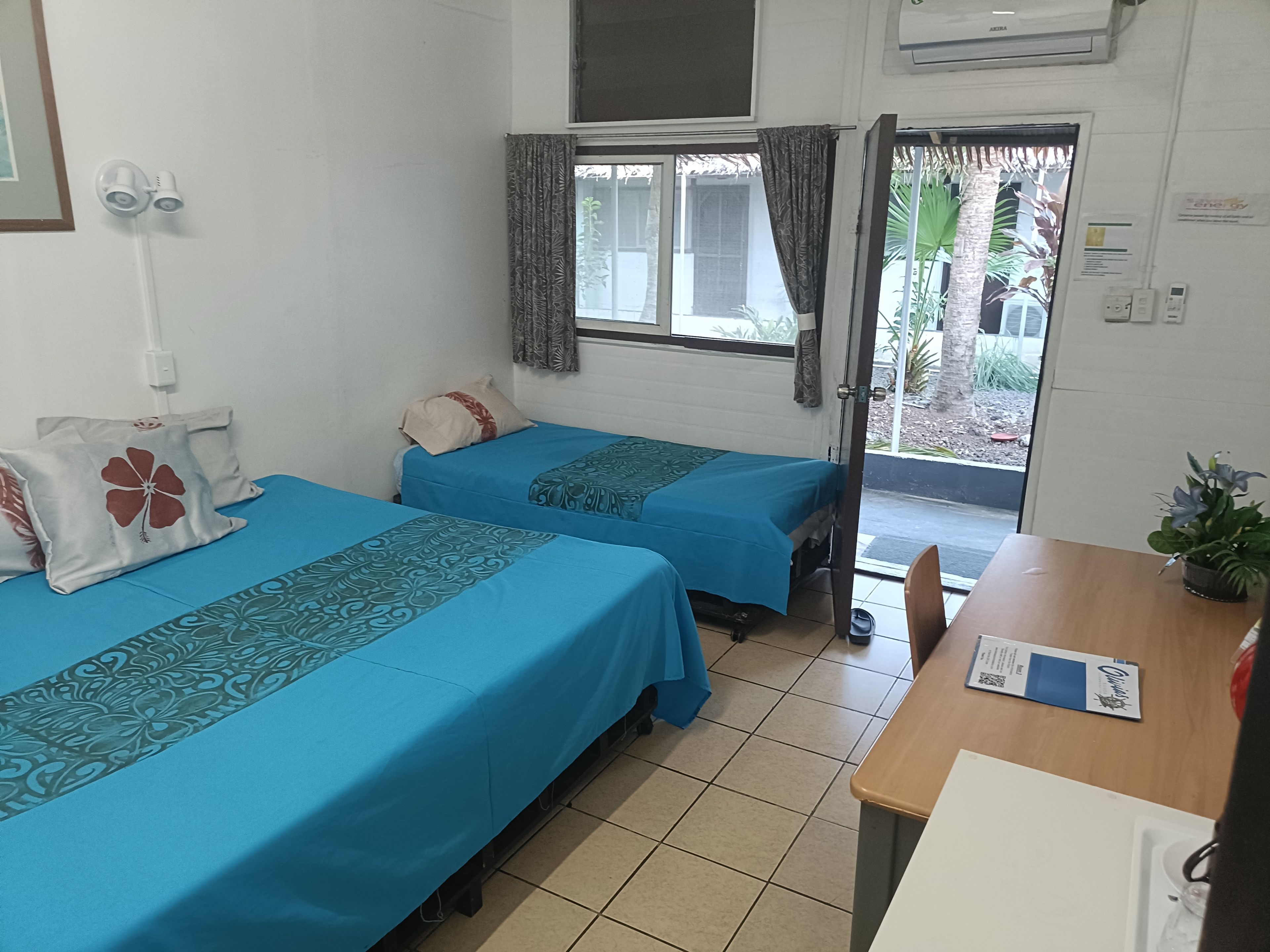 A room with two beds, a wooden table, a window with curtains, and an air conditioning unit.