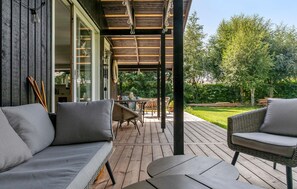 Een overdekt terras met een houten dek, rieten meubels en uitzicht op een weelderige tuin.