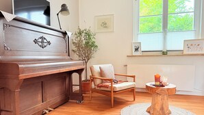 Ein Zimmer mit einem Holzpiano, einem Holzstuhl, einem kleinen Holztisch mit Kerzen, einer Topfpflanze und einem Fenster mit Jalousien.