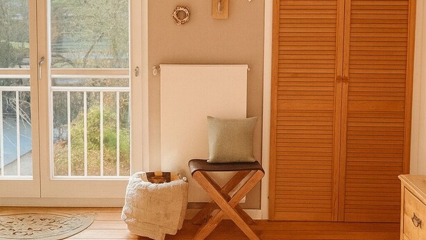 Ein Zimmer mit Holzboden, einem großen Fenster, einer Deckenleuchte, einem Holzschrank, einem kleinen Tisch mit Kissen und Wanddekorationen.