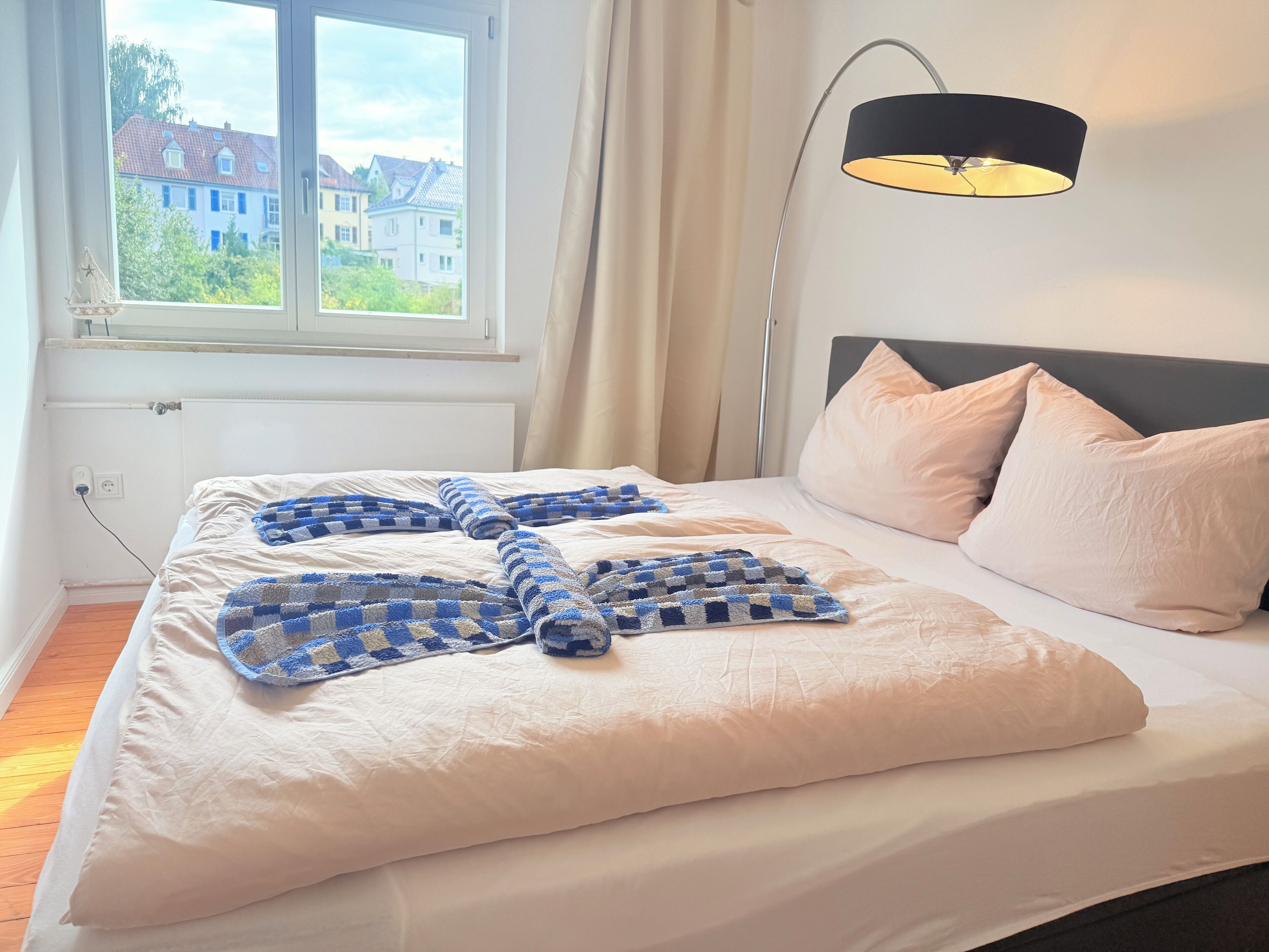 A neatly made bed with a checkered blanket, a bedside lamp, and a view of buildings through a window.
