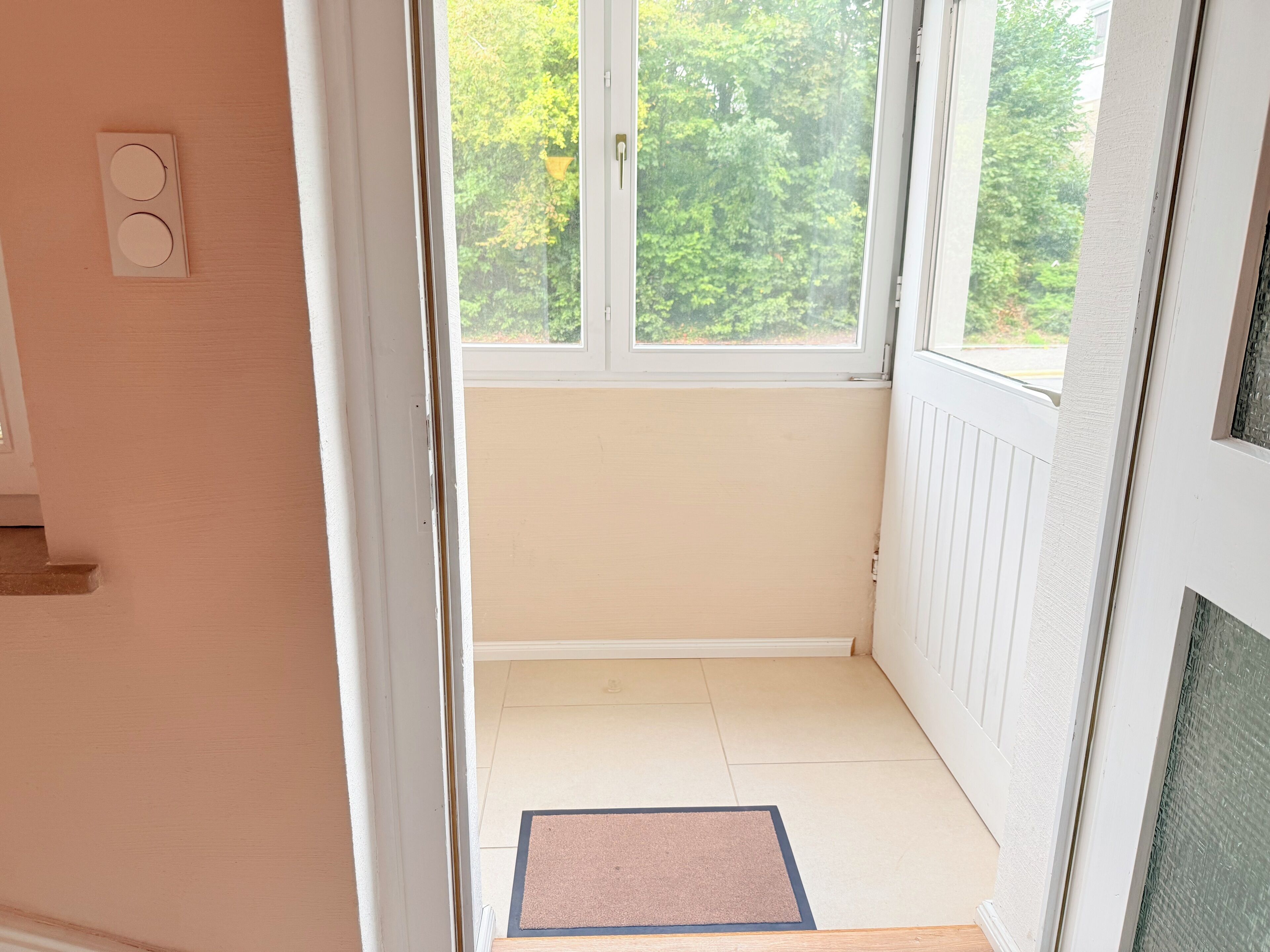 Ein Zimmer mit einer zur Balkonterrasse führenden Tür, einem Fenster mit Blick auf Grünflächen und einer Fußmatte auf dem Boden.