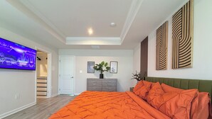 A bedroom with a large bed, a television on the wall, a dresser, and a door leading to another room.