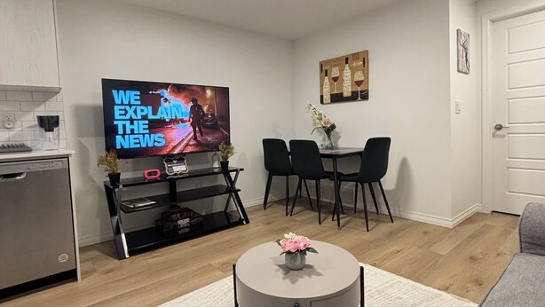 A modern living room with a flat-screen TV, a dining table with chairs, a coffee table with flowers, and a kitchen area with a dishwasher. - Chezeey’s Home - Refresh, Relax, Work & Unwind - new beautiful basement suite (Edmonton)