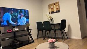 A living room with a TV, a dining table with chairs, a small table with a vase of flowers, and a shelf with electronic devices. - Chezeey’s Home - Refresh, Relax, Work & Unwind - new beautiful basement suite (Edmonton)