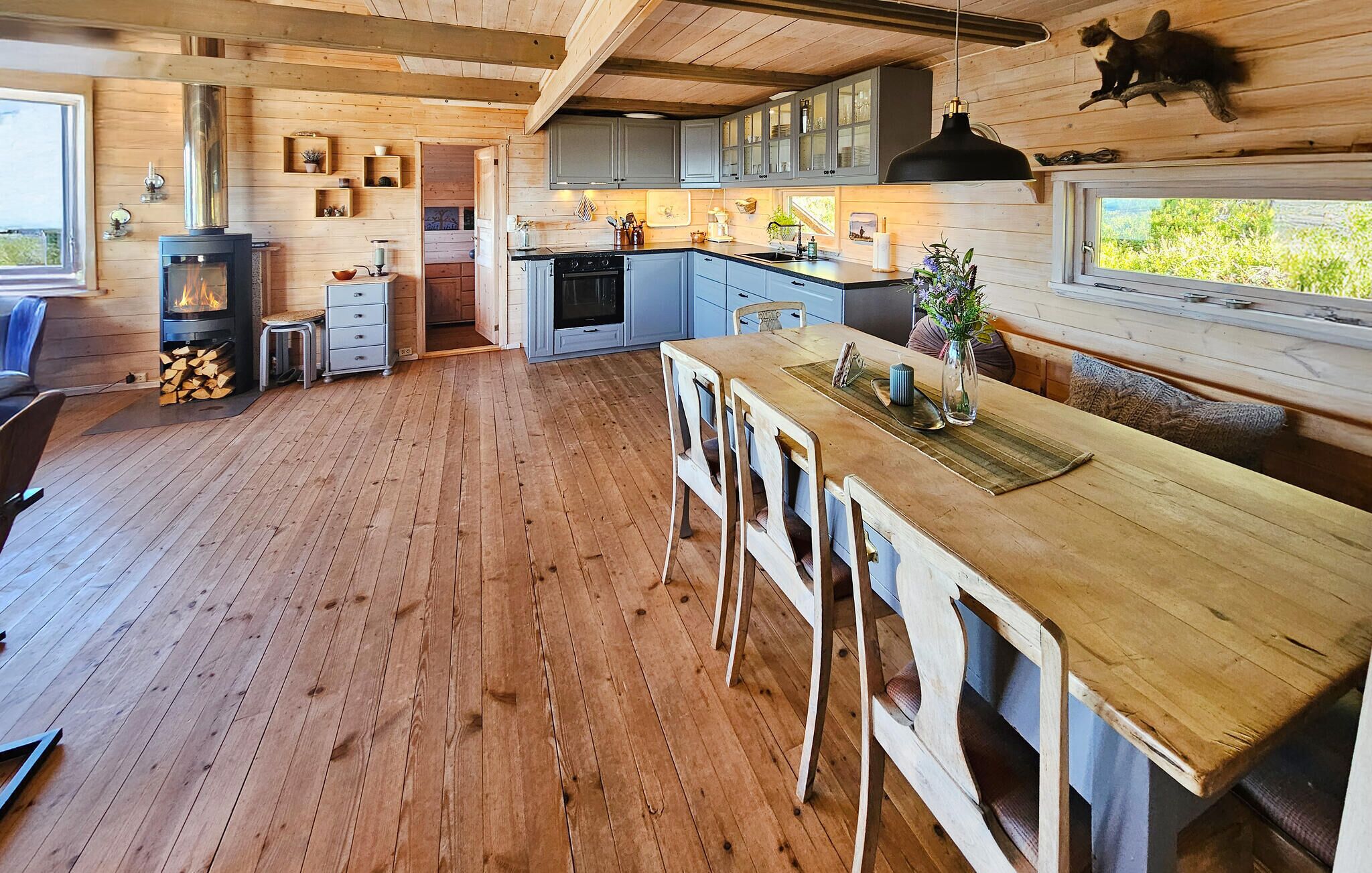 Un intérieur de chalet chaleureux avec un plancher en bois, une cuisine centrale dotée de meubles gris, un coin repas avec une table et des chaises en bois, et une cheminée.