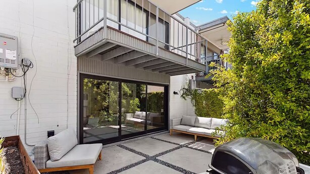 En moderne udendørs terrasse med integreret grill, en glas skyde dør der fører til en balkon, og et siddeområde med hynder.