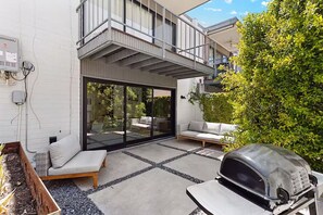 Une terrasse extérieure moderne, dotée d’un barbecue intégré, d’une porte-fenêtre coulissante donnant sur un balcon et d’un espace salon avec des coussins.