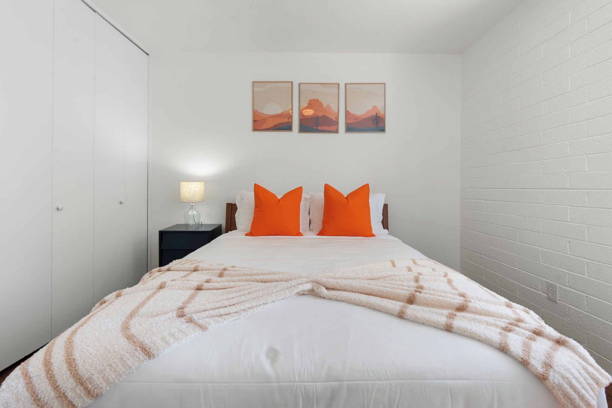 A single bed with a white duvet and orange pillows, a bedside table with a lamp, and three landscape paintings on the wall.