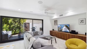 En moderne stue med en grå hjørnesofa, en gul lænestol, et fladskærms-tv og en skydedør, der fører ud til en terrasse med havemøbler.