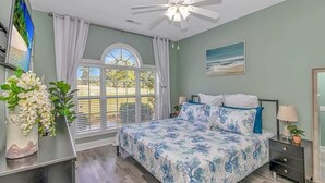 Un dormitorio con cama, ventilador de techo, ventana con persianas, un cuadro en la pared y una planta en la mesita de noche.