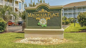 Señal de Magnolia Place con un emblema decorativo, rodeada de césped cuidado y un área de estacionamiento.