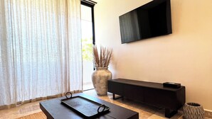 A modern living room with a dark wooden coffee table, a large vase with dried plants, a black bench, and a flat-screen TV mounted on the wall. - Peregrina Tulum 302B 1BR Condo (Tulum)