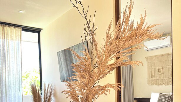 A dining table with a vase of dried plants, a sofa, and a bed in the background. - Peregrina Tulum 302B 1BR Condo (Tulum)