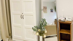 Una habitación con un armario blanco, un espejo, una pequeña mesa de madera con un teléfono y un router, y un florero con flores.
