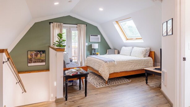 Een slaapkamer met schuine balken, een bed, een bureau, een stoel, een raam en een plant.