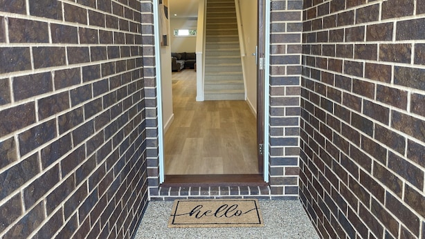 Un pasillo con paredes de ladrillo, piso de madera y una puerta con un felpudo que dice 'hello'.