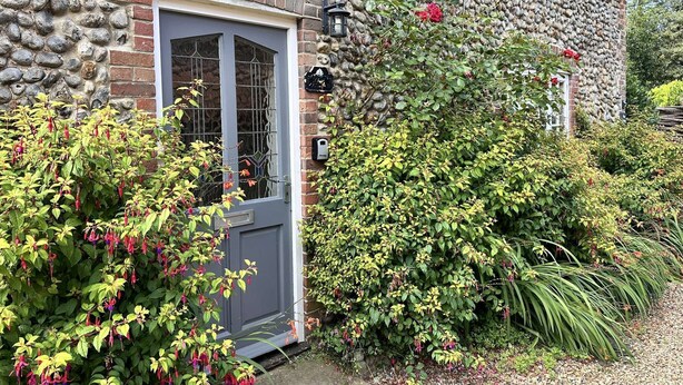 A stone house with a grey door, a small window, and a wall-mounted light fixture. - A charming detached period cottage tucked into the town of Blakeney (Blakeney)