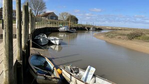 A calm river with boats docked along the shore, a wooden pier, and a building in the background. - A charming detached period cottage tucked into the town of Blakeney (Blakeney)