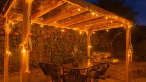 Une terrasse extérieure dotée d’une pergola en bois, de guirlandes lumineuses, d’un barbecue et de chaises en osier.