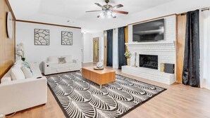 Un salon moderne avec une cheminée blanche, un ventilateur de plafond et un tapis à motifs.